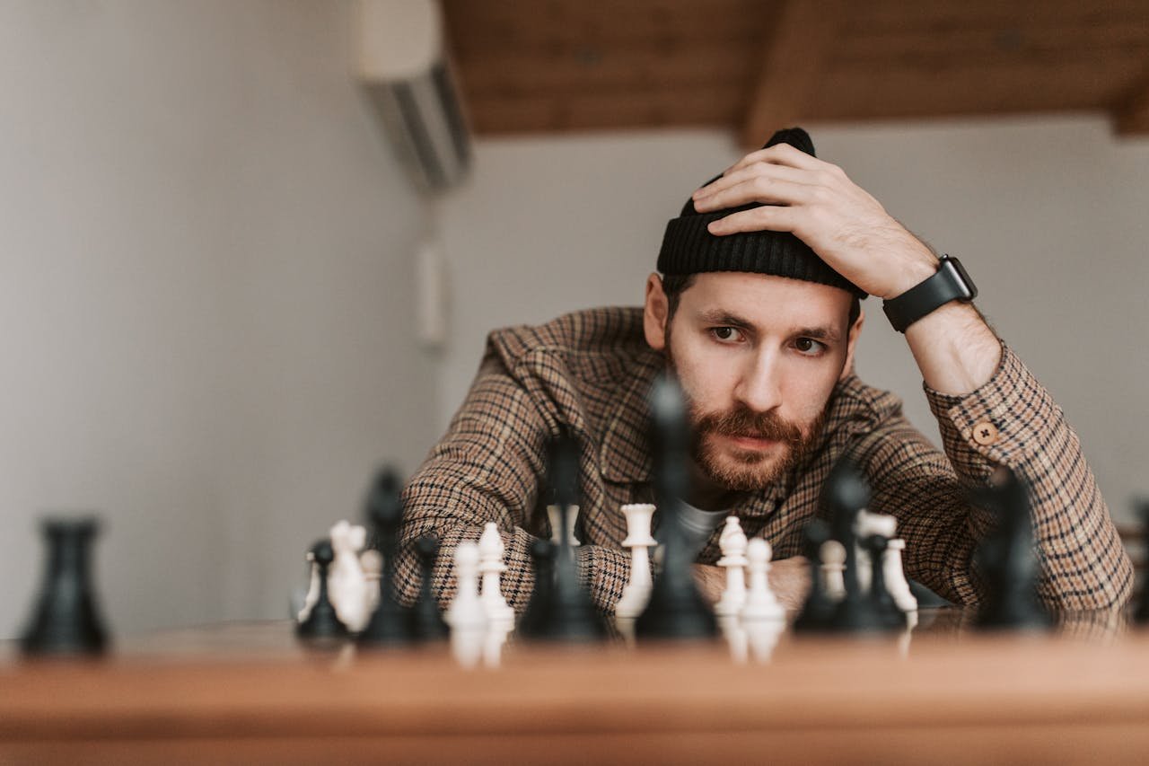 Man in checkered shirt and beanie intensely focuses on chess game indoors, showcasing strategic thinking.